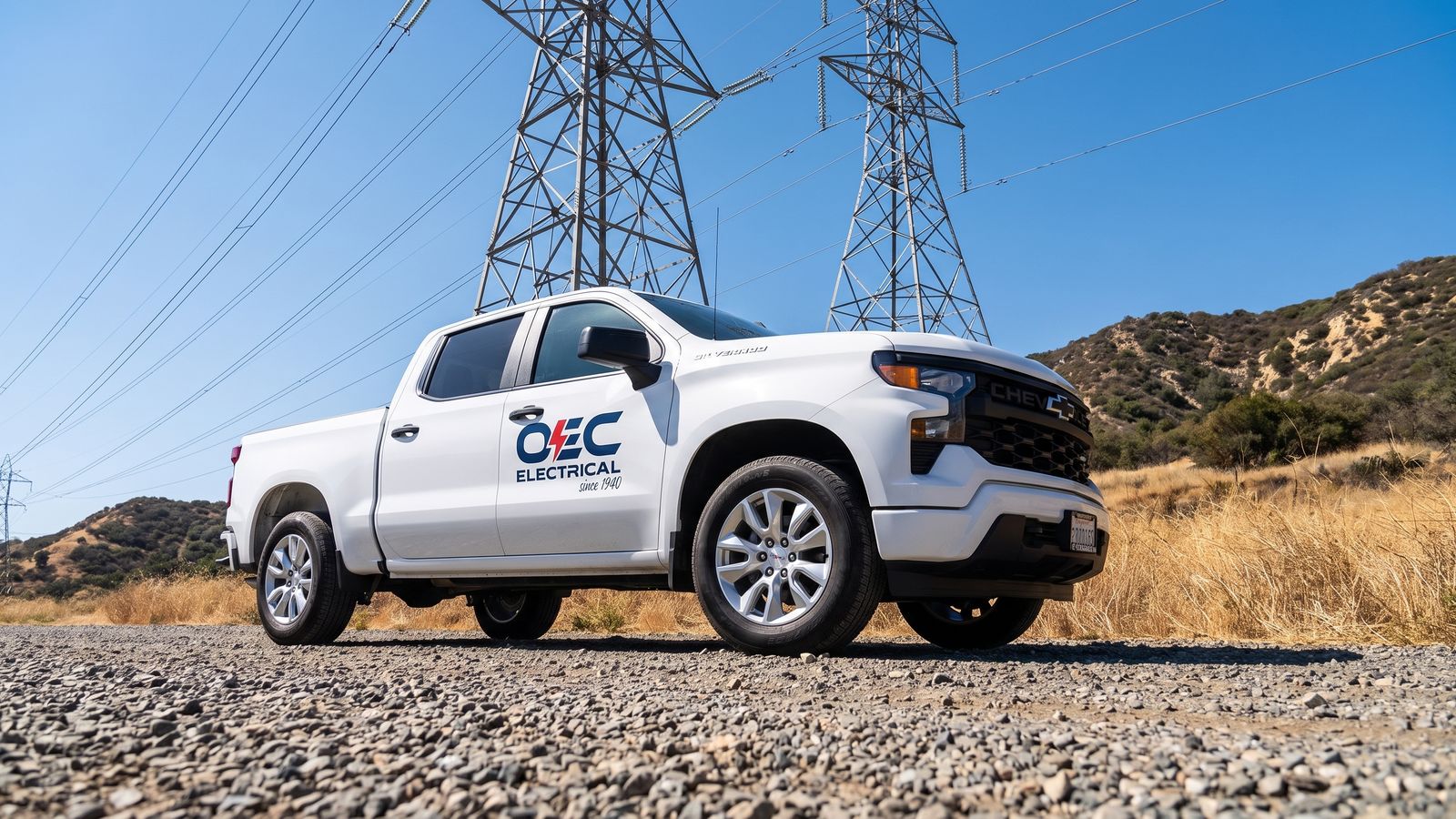 White OEC Electrical Chevrolet Silverado service truck parked beneath steel high-voltage transmission towers